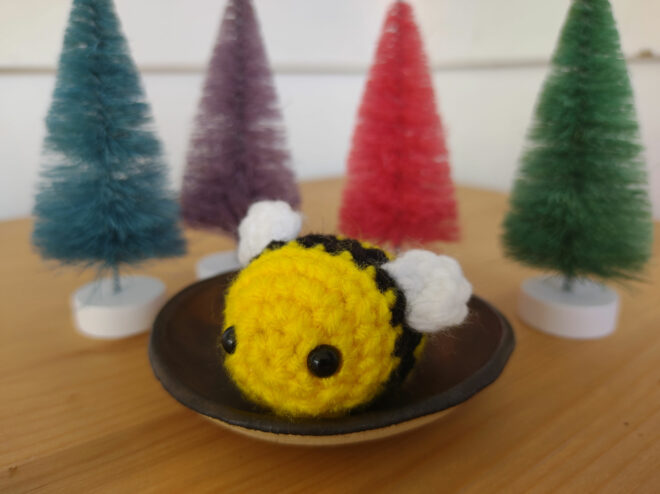 Crochet bee sits in front of tiny multicolored pine trees